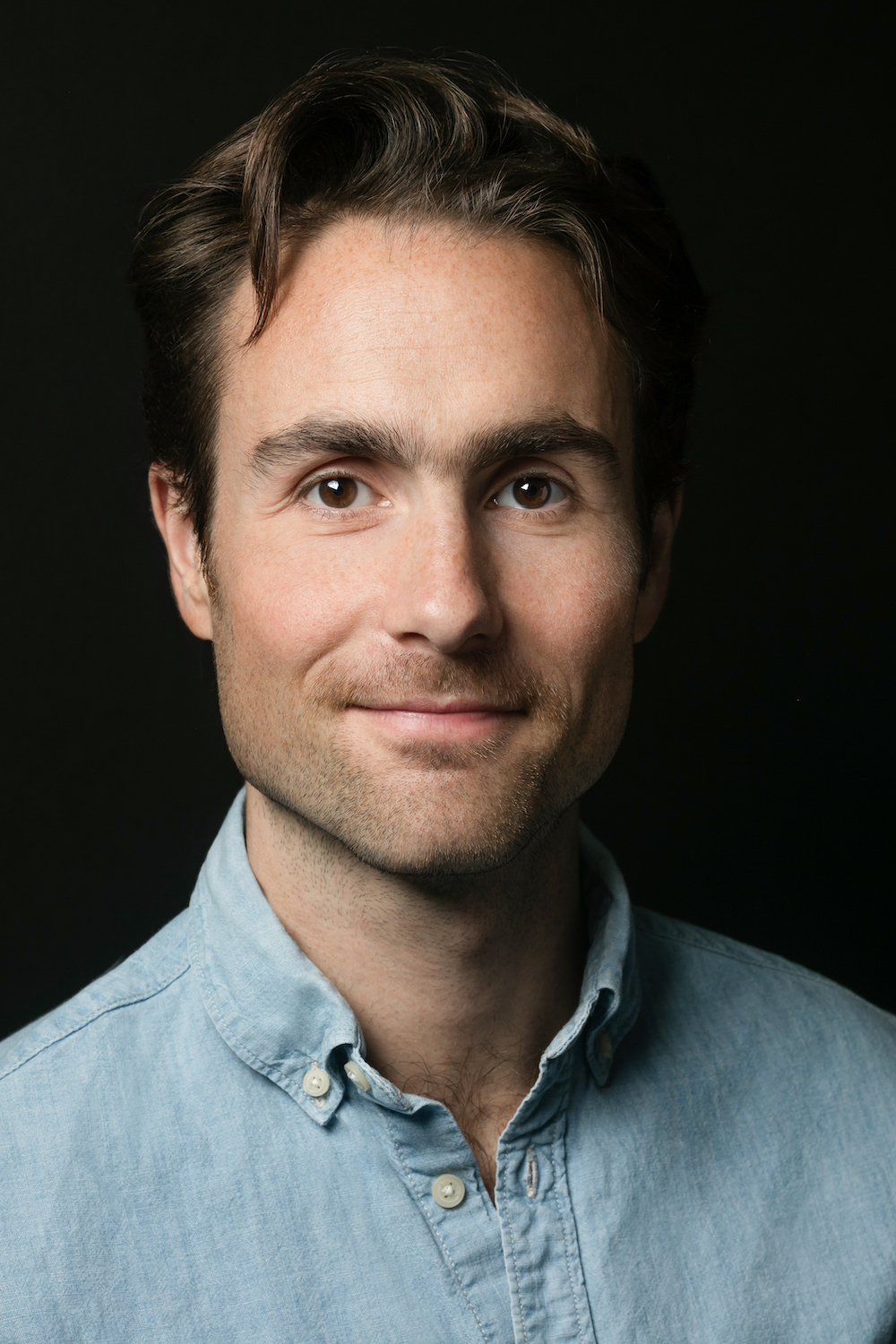 David Wallace Wells looks into the camera with a closed mouth smile. He is a caucasian man with dark brown hair and brown eyes. He is wearing a blue button-down and is in front of a black background